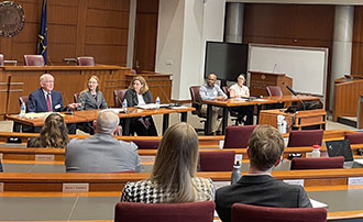 Panelists seated at the attorney tables in front of an audience in the court room